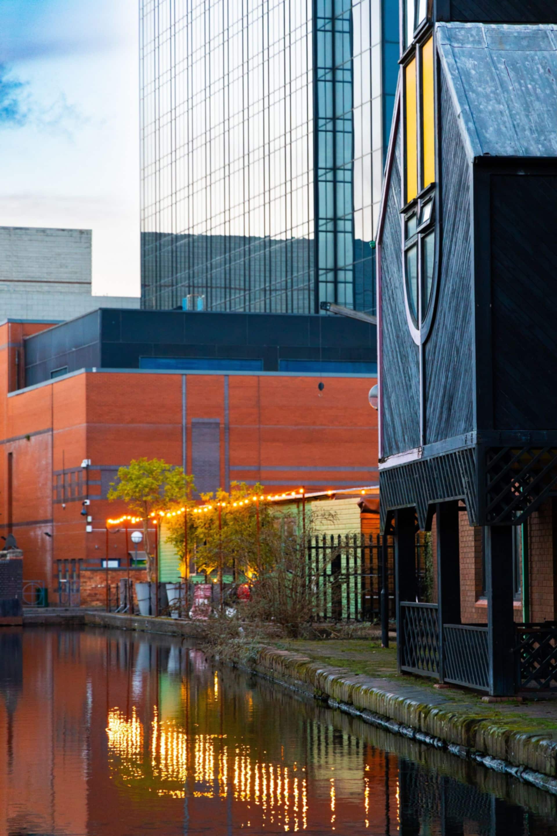 String lights reflected in a Birmingham canal at dusk beside red-brick buildings.