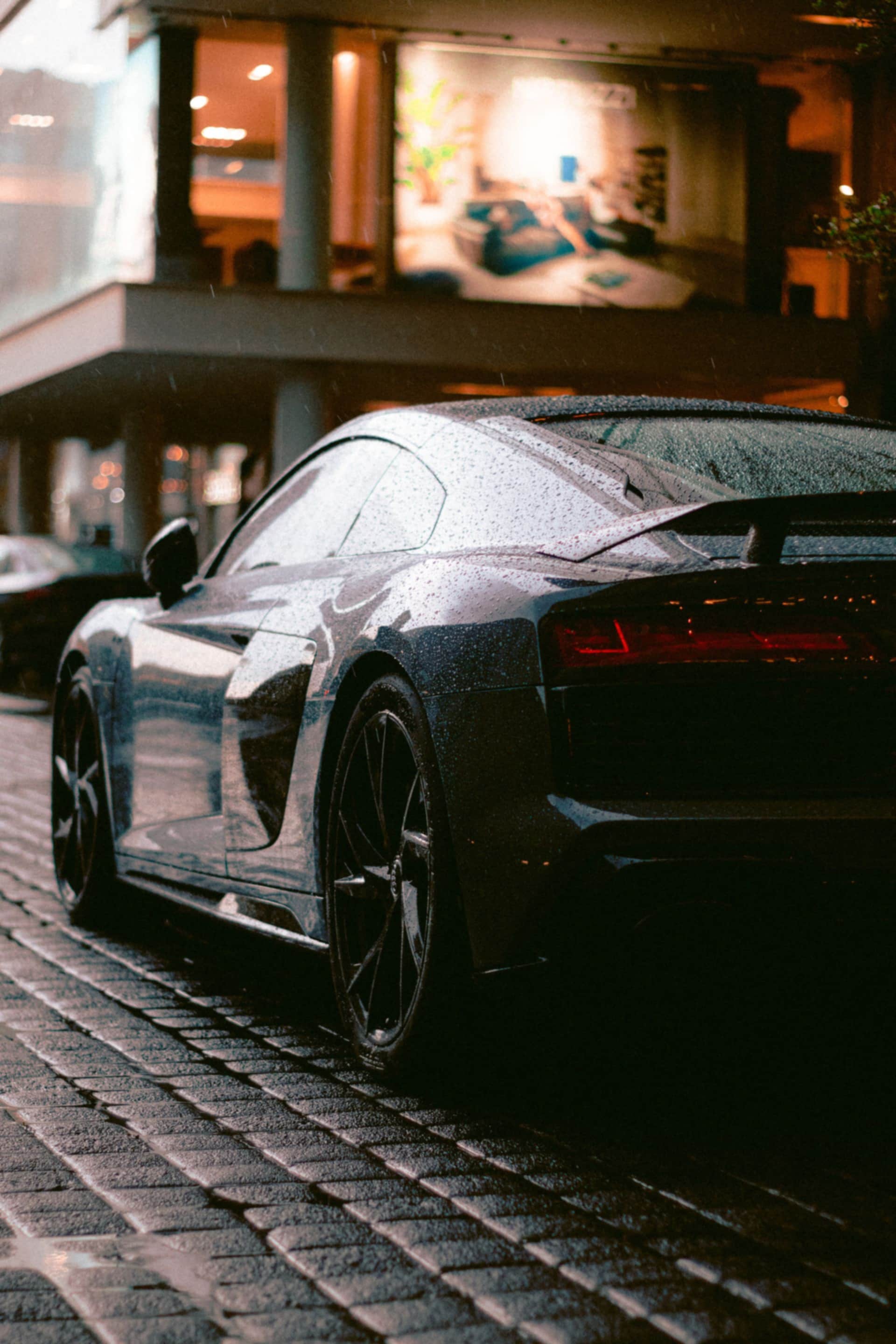 The rear three-quarter of a dark car on wet cobbles at night, warm shop light behind.