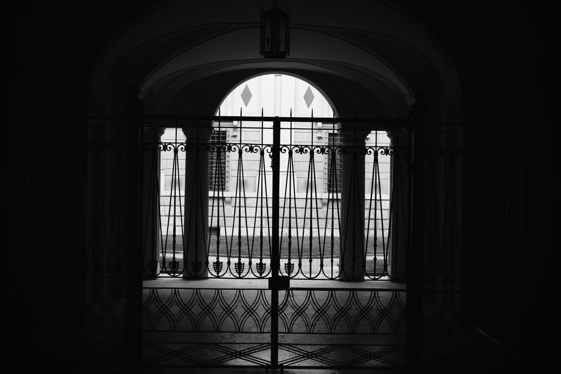An ornate wrought-iron gate at night, light in the arched windows beyond.