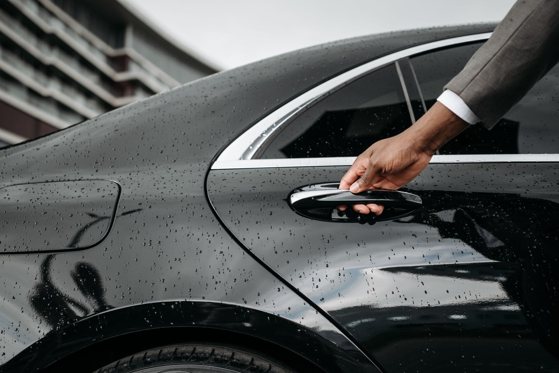 A hand reaching for the door handle of a black car in the rain.