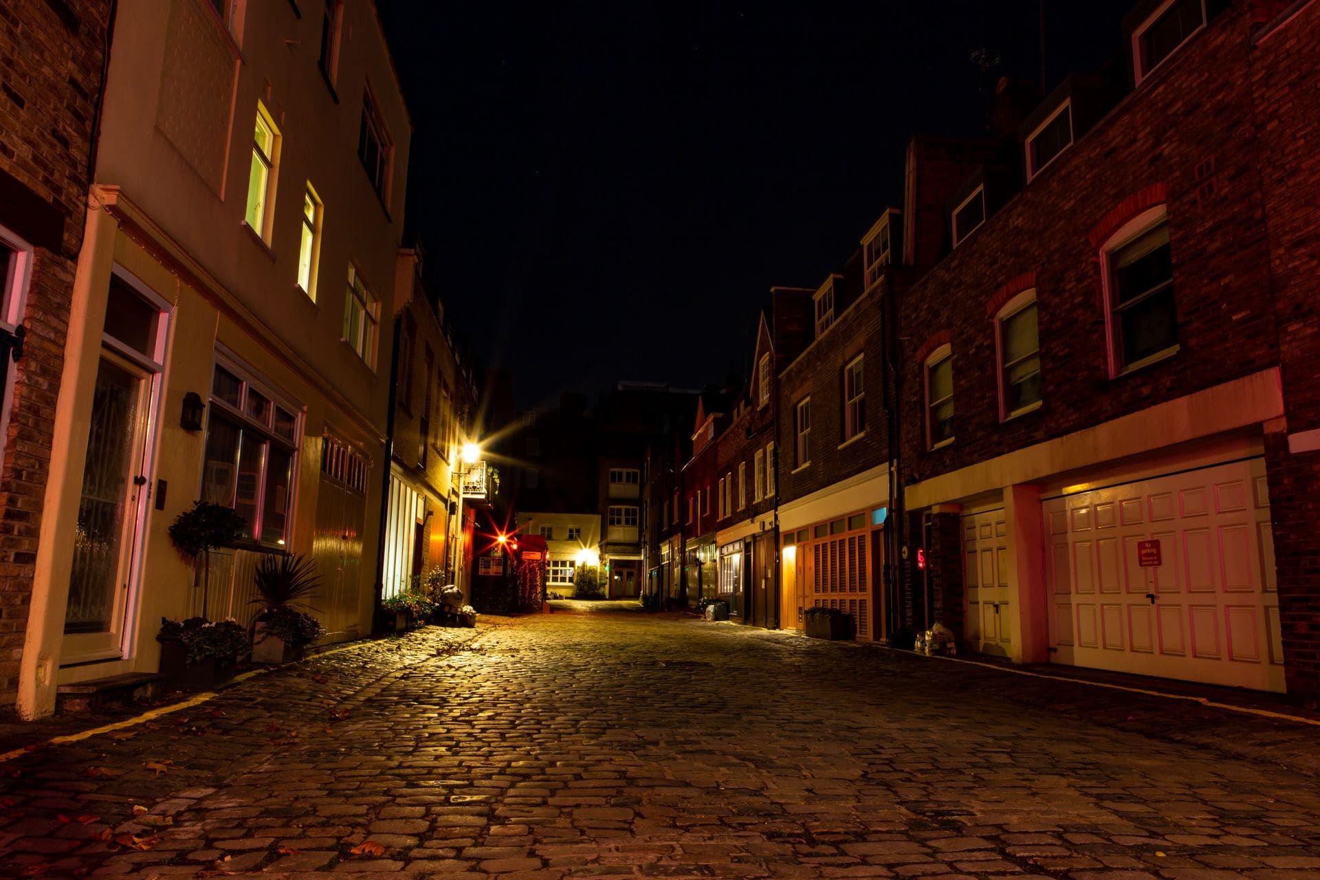 Cobbles and garage doors of a London mews lit by lamps at night.