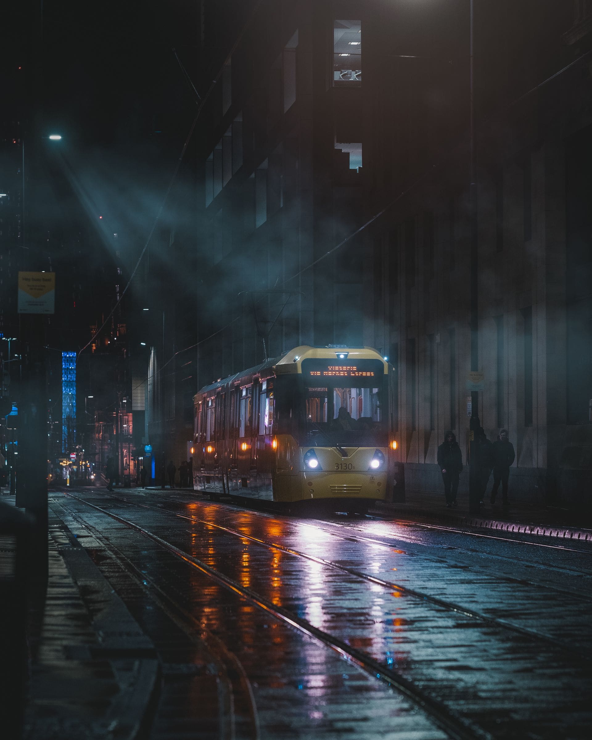 A Manchester tram crossing a wet street at night in light haze.