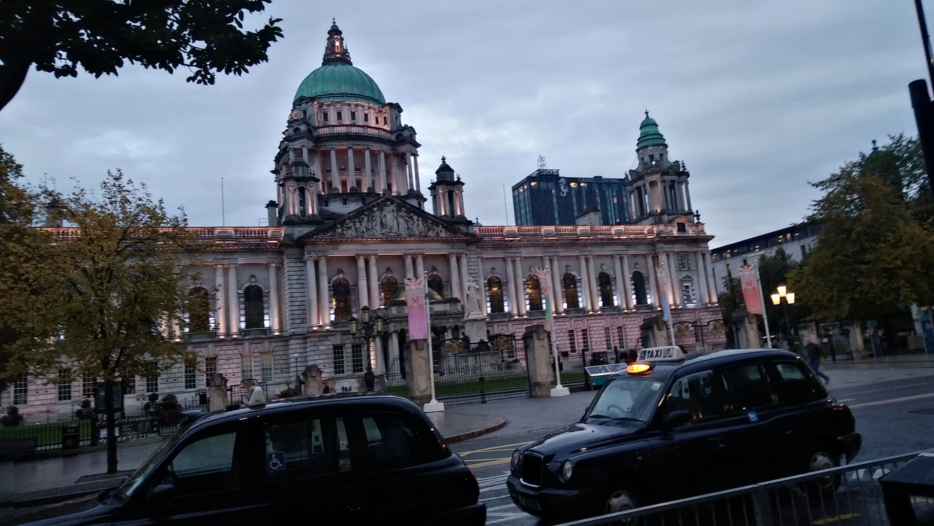 Belfast City Hall's dome and floodlit facade at dusk, a black cab passing.
