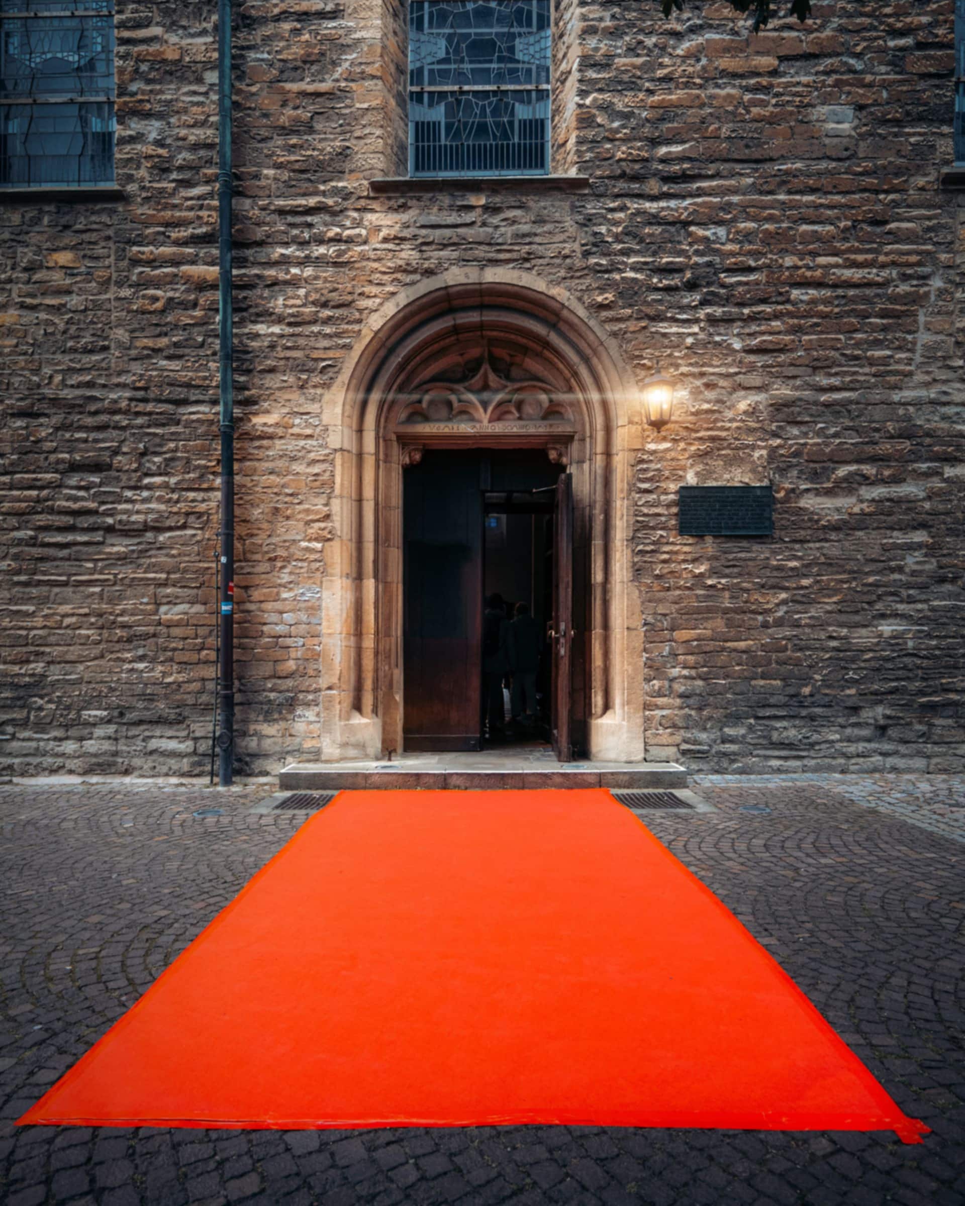 An empty red carpet leading across cobbles to a lamp-lit stone doorway in the evening.